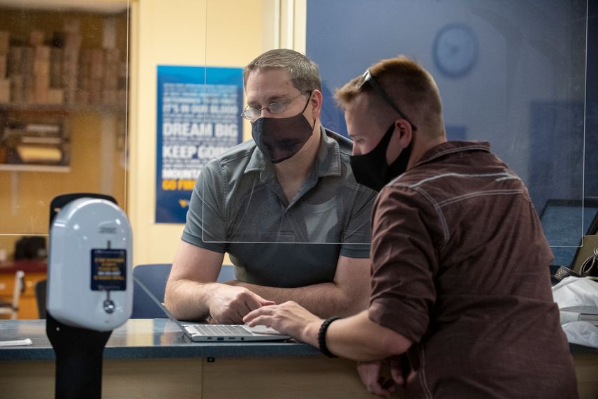 A WVU staff member, behind plexiglass and wearing a mask, assists a WVU student, also wearing a mask.